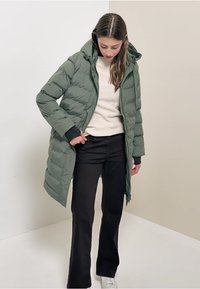 Kvinde justerer sin lynlås grønne puffjakke med hætte, iført en beige sweater, sorte bukser og hvide sneakers mod en ensfarvet lys baggrund.