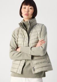 Kvinne med kort mørkt hår iført en lys beige polstret vest over en ribbestrikket høye hals-genser, med armene krysset, foran en ensfarget lys bakgrunn.