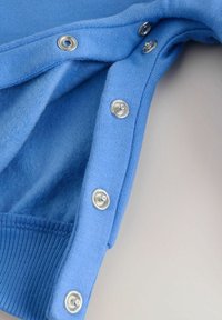 Svetlo modra pulover z mehko teksturo, z srebrnimi sponkami ob robu in rebrastim vzorcem na spodnjem robu.