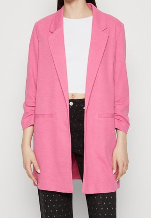 Vrouw draagt een felroze op maat gemaakte blazer over een witte crop top en zwarte broek met sterrenpatroon, staand tegen een effen achtergrond.