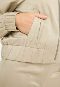 Giacca bomber beige chiaro con una texture satinata liscia. Presenta polsini e vita elasticizzati, una tasca laterale e dettagli con zip argentata.