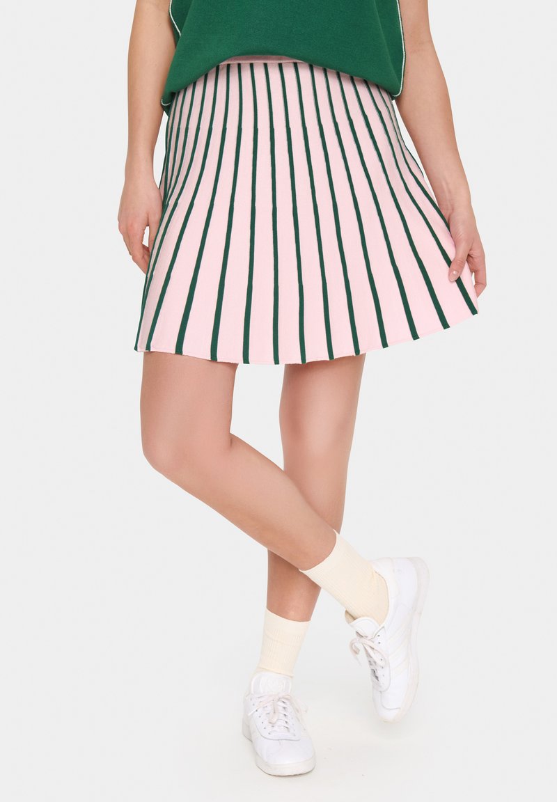Jupe plissée avec des rayures verticales roses et vertes, réalisée en tissu léger. Associée à des baskets blanches et des chaussettes beige clair.