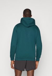 Felpa con cappuccio color verde scuro, dotata di tasca a marsupio, cappuccio regolabile, polsini e orlo a coste. Indossata con pantaloni corti grigi, che mostrano una texture liscia.