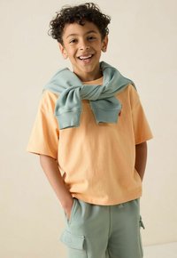 T-shirt en coton orange clair à manches courtes, associé à un pantalon cargo vert menthe. Un pull vert menthe assorti est drapé autour du cou.