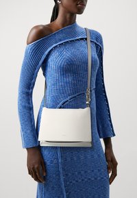 Borsa a tracolla bianca in pelle con superficie liscia, design minimalista e dettagli in metallo argentato. Indossata con un vestito blu a coste.