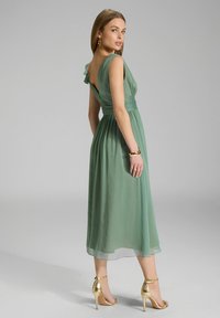 Grünes, fließendes Midi-Kleid aus leichtem Stoff, mit Rüschendetail an der Schulter, taillierter Taille und transparentem Saum. Getragen mit goldenen High Heels.