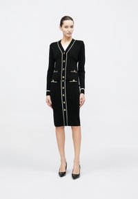 Μαύρο φόρεμα με δομημένο σχέδιο, λευκή διακόσμηση, χρυσά κουμπιά και εφαρμοστή σιλουέτα. Μακριά μανίκια και μήκος μέχρι το γόνατο.