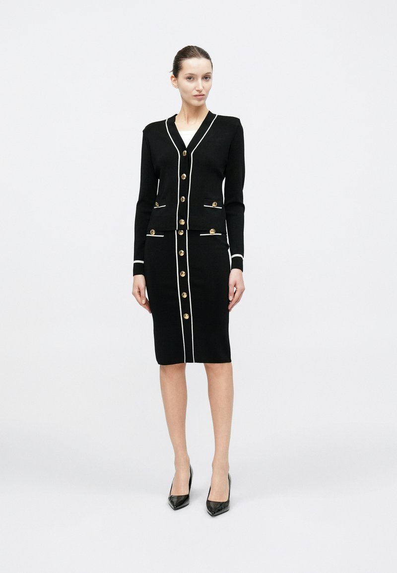 Μαύρο φόρεμα με δομημένο σχέδιο, λευκή διακόσμηση, χρυσά κουμπιά και εφαρμοστή σιλουέτα. Μακριά μανίκια και μήκος μέχρι το γόνατο.