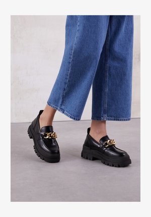 Black leather loafers with a chunky sole and gold chain detail, paired with wide-leg blue denim trousers. Textured surface, minimal design.