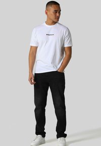 White cotton t-shirt with a black logo on the chest, paired with dark denim jeans and white sneakers, featuring a fitted cut and short sleeves.