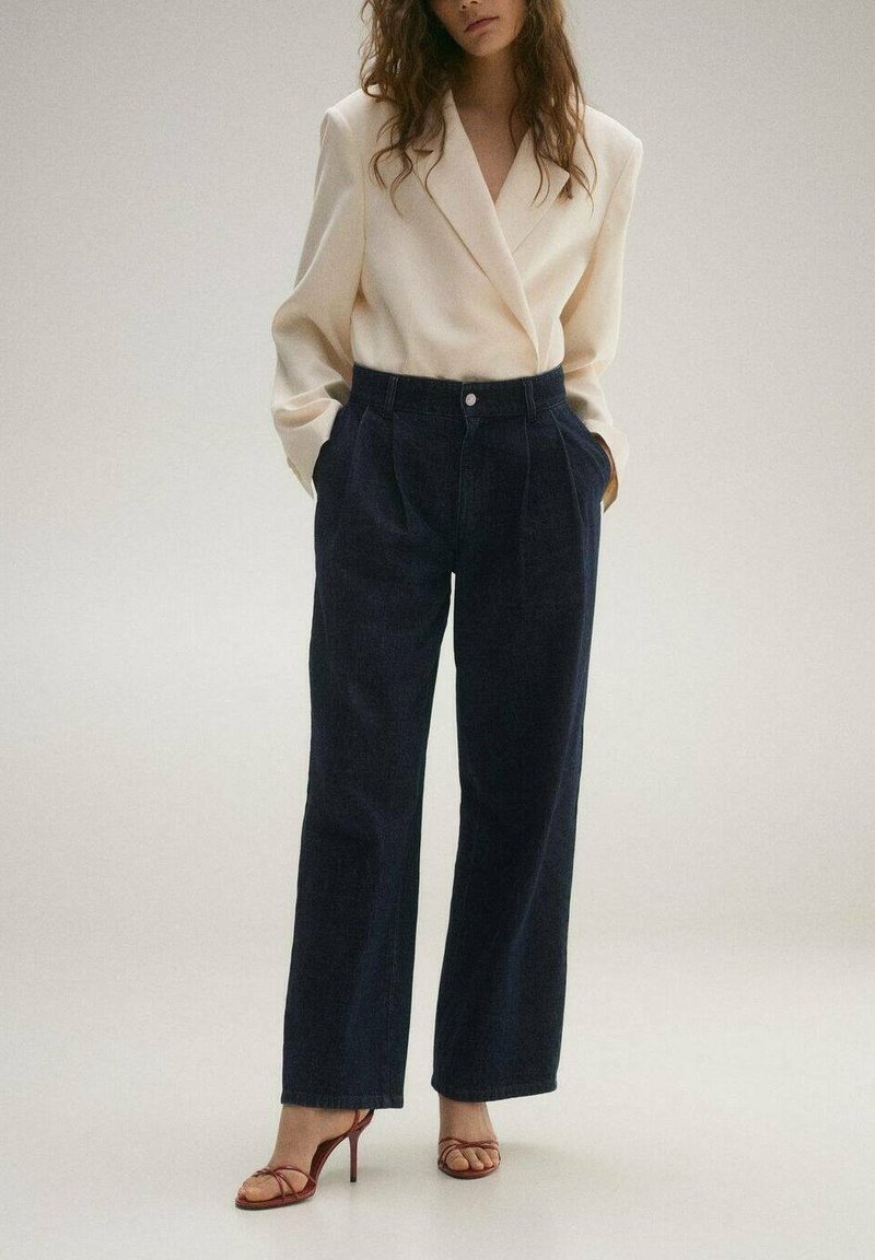 Krem blazer z ovitim dizajnom, v kombinaciji s temno modrimi hlačami širokega kroja. Model nosi rjave sandale s peto. Nežen, teksturiran material.