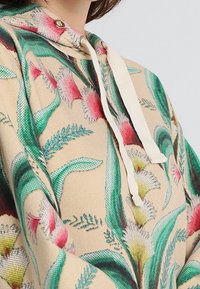 Huvtröja i beige med ett blommönster som har gröna blad och rosa inslag, tillverkad av strukturerad tyg, med krämfärgade dragsko.