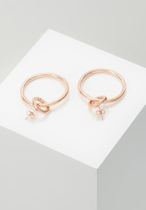 Paire de boucles d'oreilles créoles en or rose avec un petit détail en forme de nœud et des attaches à tige, présentées sur un socle blanc.