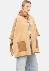 Vrouw draagt een beige cape met bruin doorgestikte zakken en een capuchon, gecombineerd met lichtblauwe jeans met wijde pijpen en een wit topje.