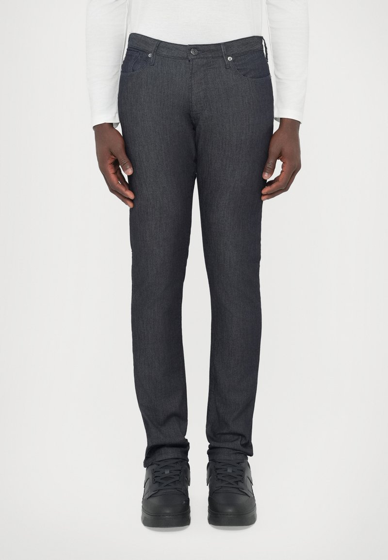 Σκούρο τζιν παντελόνι με στενή εφαρμογή, σχεδίασ�η με πέντε τσέπες και διακριτή υφή. Συνδυάζεται με μαύρα ψηλά αθλητικά παπούτσια.