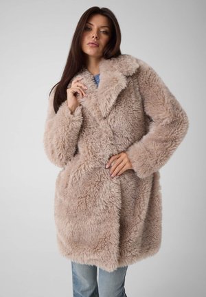 Une femme porte un long manteau beige moelleux sur un jean et en tient le col devant un fond de studio gris uni.