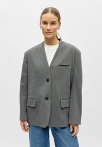 Veste en laine mélangée grise avec un col cranté, deux poches avant, une poche poitrine unique et deux boutons noirs. Portée sur une chemise blanche.