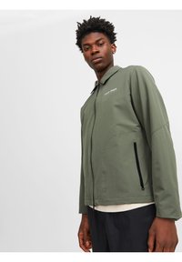 Veste olive légère avec fermeture éclair sur le devant, poches latérales et un logo subtil. Texture lisse et design moderne, minimaliste.
