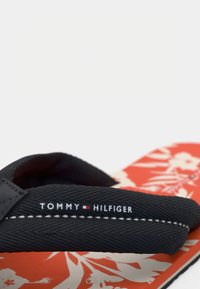 Black fabric flip-flop strap with Tommy Hilfiger logo over orange floral-patterned sole.