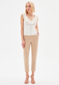 Valkoinen laskostettu tanktoppi, jossa on struktuuripinta, yhdistettynä beigekuituneisiin housuihin. Asusteessa on herkkiä pitsidetails ja slim fit -malli.