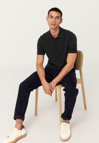 Donkergroene polo met een kraag, gecombineerd met donkerblauwe broek en witte sneakers. Model zittend op een houten stoel.