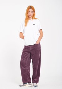 Camiseta de algodón blanca con mangas cortas y logo negro, combinada con pantalones de pana morada de pierna ancha y texturizada, y zapatillas multicolor.