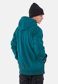 Man met een teal gekleurde capuchonjas met de tekst "SLADE" op de onderrug en een zwarte muts, handen in zakken, kijkt naar rechts tegen een witte achtergrond.