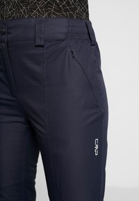 Mørkeblå udendørsbukser lavet af elastisk materiale med en lynlåslomme på siden og et CMP-logo på låret. Glat tekstur.