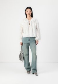 Blusa branca transparente com textura floral, decote redondo e cordão. Combinada com jeans de perna larga azul claro e uma pequena mala cinza.