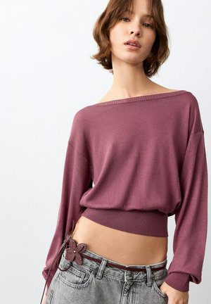 Giovane donna che indossa un maglione mauve con spalle scoperte e jeans grigi scoloriti con una cintura decorativa a fiore, posa su uno sfondo bianco.