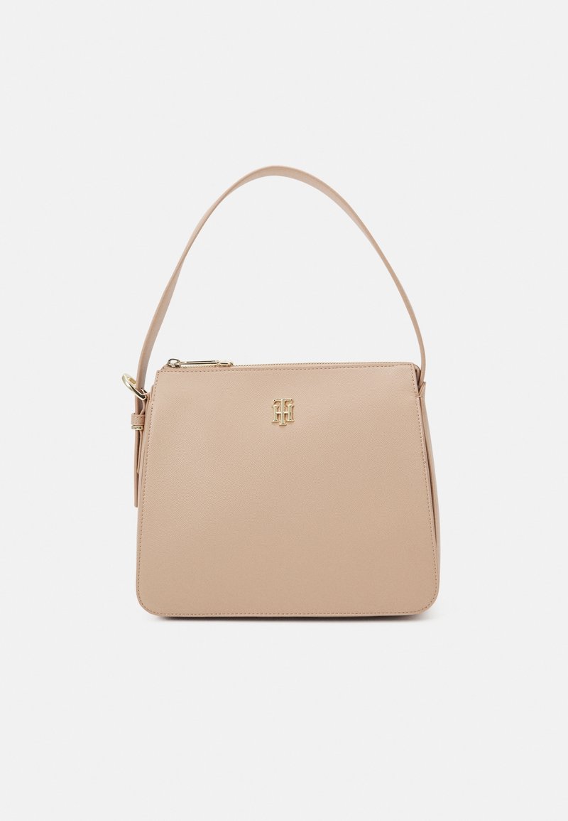 Sac à main structuré beige avec une courte anse, une fermeture éclair dorée et un petit logo doré sur le devant, posé sur un fond blanc uni.