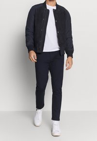 Chaqueta bomber azul marino con parte delantera de ante y mangas acolchadas, llevada sobre una camiseta blanca, combinada con pantalones azul marino de corte ajustado y zapatillas blancas.