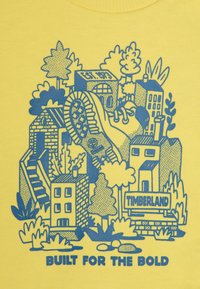 Žlté tričko s modrou grafickou ilustráciou budov, stromov a stopy topánky, s textom "TIMBERLAND" na spodnej strane.
