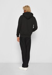 Sort hættejakke med kengurulomme og snøre i hætten. Har ribbede manchetter og kant. Bæres med løse sorte bukser og hvide sneakers.