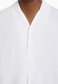 Camicia bianca a maniche corte realizzata in tessuto leggero, con colletto a punta e cinque bottoni sul davanti. Texture liscia.