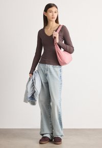 Top ajustat cu mânecă lungă de culoare maro, blugi de culoare bleu deschis cu picior larg, geantă de umăr roz și adidași maro. Jachetă aruncată pe braț. Fundal simplu.