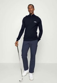 Jūras spēku zips galds golfam ar mīkstu audumu un pieguļošu dizainu. Apvienots ar jūras spēku biksēm un baltām golfu kurpēm, tur rokā golfu nūju.
