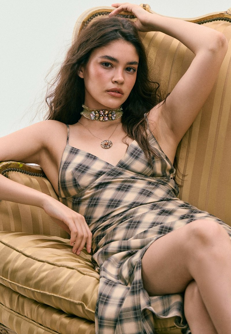 Jeune femme aux longs cheveux portant une robe à carreaux et un collier orné de bijoux, s'allongeant sur un fauteuil rayé et rembourré.
