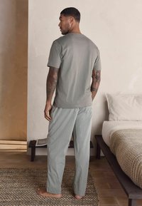 Graues Baumwoll-T-Shirt und hellgrüne gestreifte Schlafanzughose. Modell von hinten betrachtet, stehend auf einem strukturierten Teppich in einem minimalistischen Umfeld.