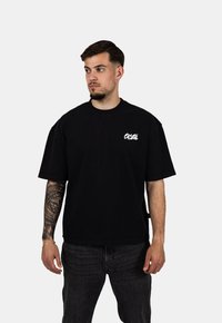 Czarna, oversized bawełniana koszulka z krótkim rękawem, z białym logo na lewej piersi. Noszona z ciemnymi dżinsami.