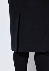Cappotto nero in misto lana con silhouette dritta, caratterizzato da uno spacco laterale e una texture liscia, abbinato a pantaloni neri aderenti.