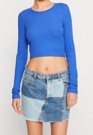 Kobieta ubrana w jaskrawo niebieski długi crop top oraz spódniczkę mini z denimu w stylu patchwork, z postrzępionym dołem.