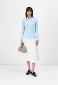 Camicia a maniche lunghe a righe blu chiaro, gonna midi bianca, scarpe nere, in mano una piccola borsa a pochette di lavanda. Tessuto morbido, vestibilità sagomata.