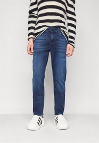 Jeans oscuros con corte slim, combinados con un suéter a rayas en blanco y negro y zapatillas blancas con rayas negras, de pie sobre un fondo liso.