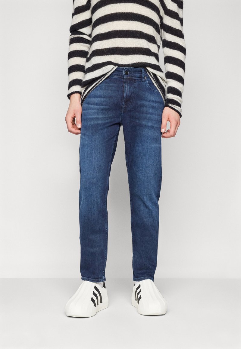 Jeans oscuros con corte slim, combinados con un suéter a rayas en blanco y negro y zapatillas blancas con rayas negras, de pie sobre un fondo liso.