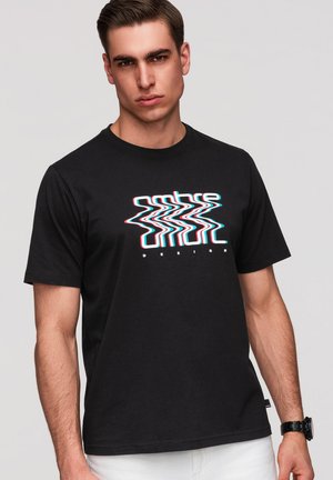Mann mit kurzem braunen Haar, der ein schwarzes T-Shirt mit welliger Textgrafik und weiße Hosen trägt, steht vor einem einfarbigen hellen Hintergrund.