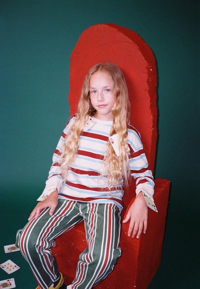 Kind zit in een rode sculpturale stoel, draagt een gestreepte lange mouwen shirt en gestreepte broek, met speelkaarten zichtbaar op de mouwen en de vloer.