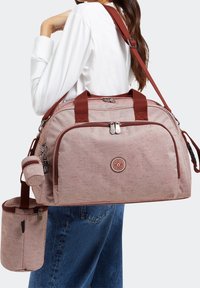 Kvinde iført hvid skjorte og blå jeans bærer en lys pink herringbone duffeltaske med maroon stropper samt en aftagelig lille pung og flaskeholder.