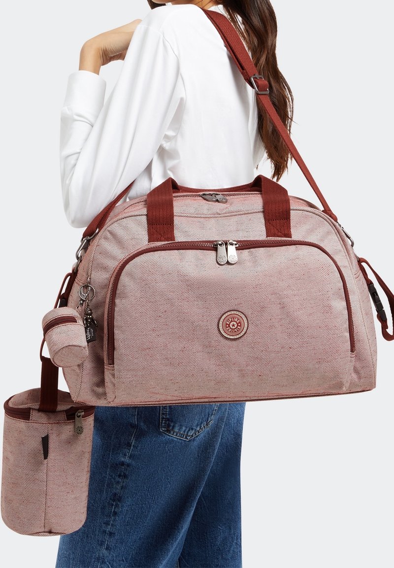 Kvinde iført hvid skjorte og blå jeans bærer en lys pink herringbone duffeltaske med maroon stropper samt en aftagelig lille pung og flaskeholder.