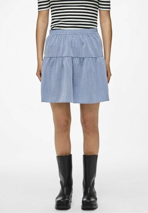 Kvinde iført en lyseblå lagdelt nederdel, sorte støvler til midt på læggen og en kortærmet sort-hvid striped top, stående mod en hvid baggrund.